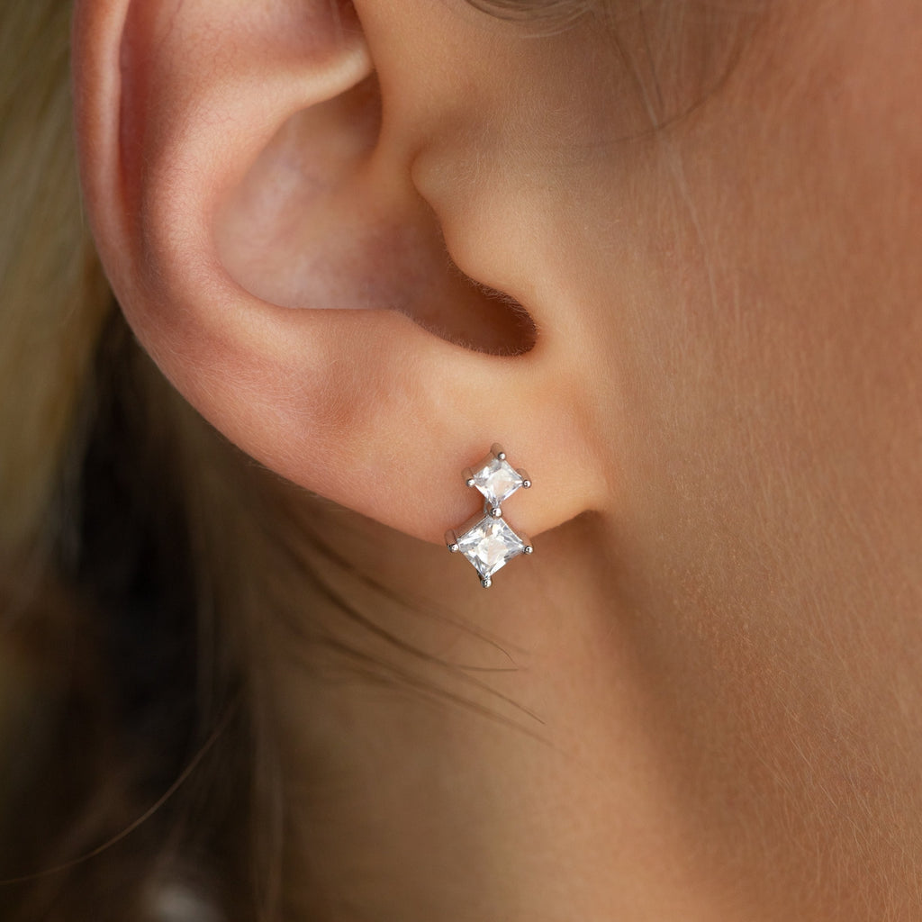 Close-up of an ear adorned with Aria Diamond Drop Studs—sleek, lightweight earrings showcasing two square, clear gemstones in a vertical setting, perfect for adding everyday elegance.
