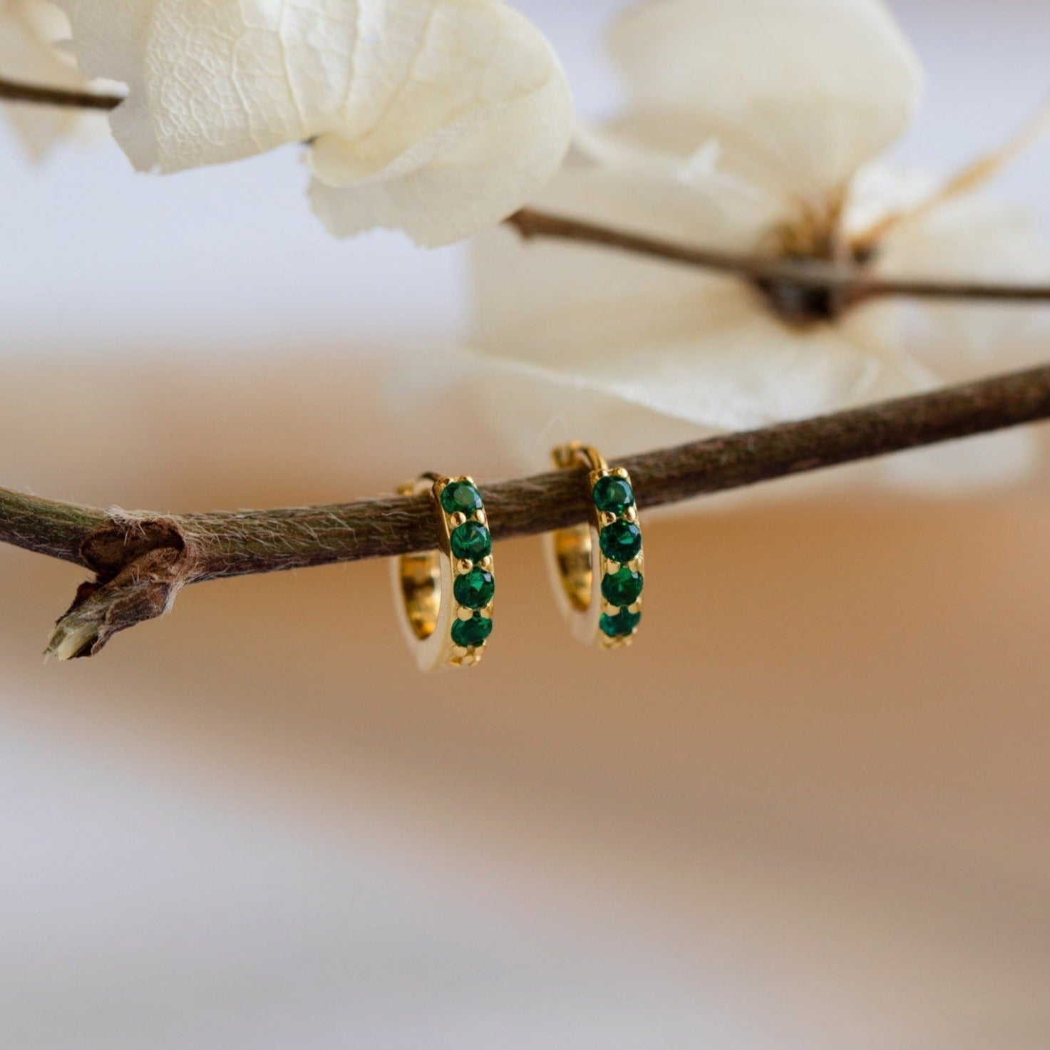 The Elise Emerald Huggies are small gold earrings with emerald stones, shown hanging on a branch near delicate white dried flowers.