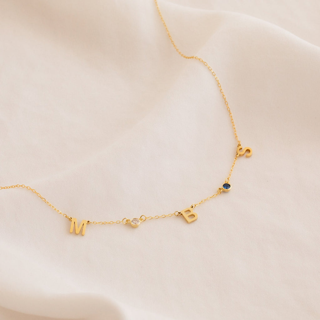 The Alice Letter Birthstone Necklace features gold M, B, and S charms with clear and blue gems on soft white fabric—an elegant, personalized gift perfect for mom.