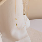 The Sideways Initial Birthstone Necklace features a gold S charm and green birthstone bead, making it a thoughtful gift for Mom. Beautifully presented on a soft white fabric background.