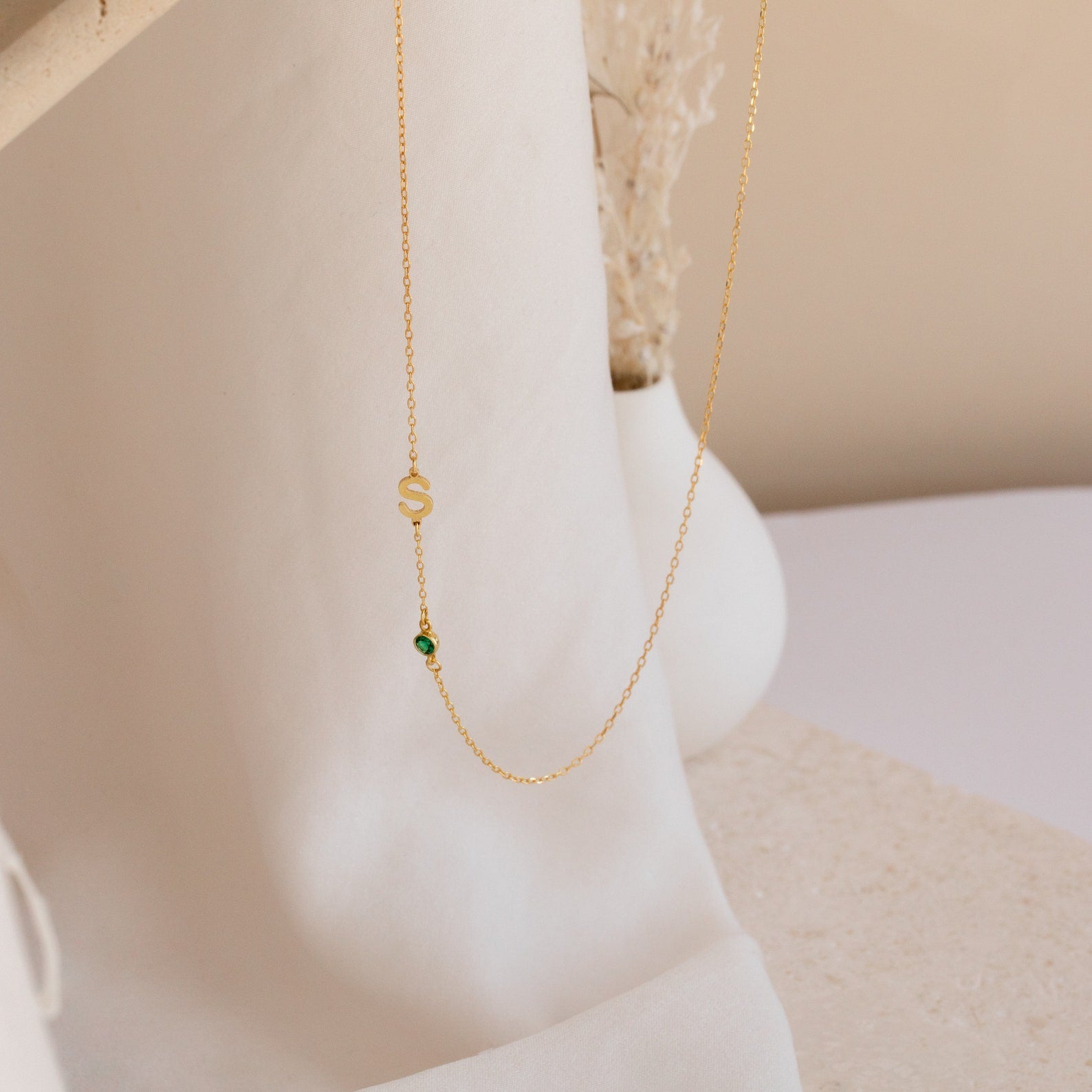The Sideways Initial Birthstone Necklace features a gold S charm and green birthstone bead, making it a thoughtful gift for Mom. Beautifully presented on a soft white fabric background.
