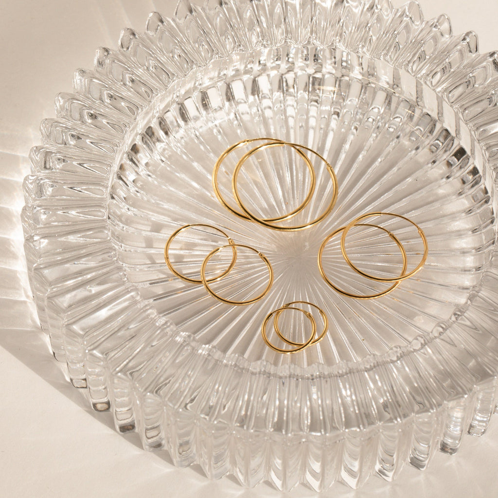 Endless Thin Hoops rest on a textured clear glass dish with sunlit shadows and reflections, highlighting these gold earrings as a timeless and versatile everyday accessory.