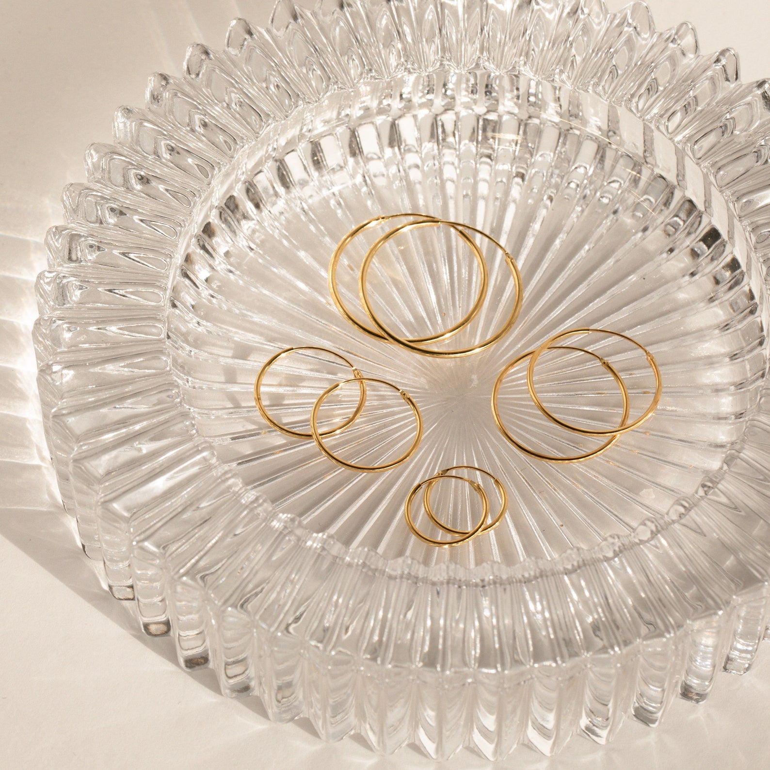 Endless Thin Hoops rest on a textured clear glass dish with sunlit shadows and reflections, highlighting these gold earrings as a timeless and versatile everyday accessory.