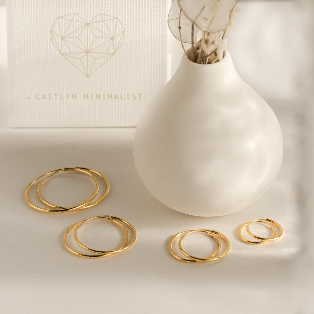 Our Endless Thin Hoops, timeless gold hoop earrings in various sizes, are shown next to a white vase and minimalist card—perfect for everyday wear and effortless style.