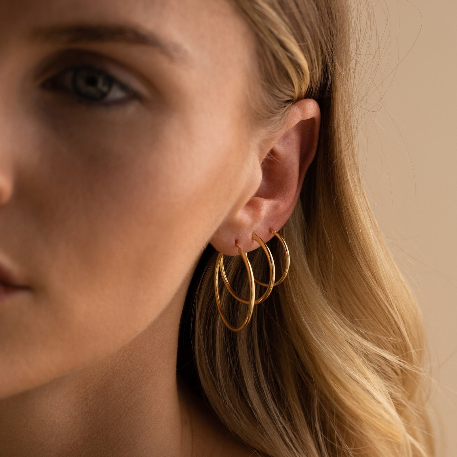 A close-up shows a woman with blonde hair wearing three overlapping Endless Thin Hoops on her ear, softly lit. These classic earrings are a stylish everyday accessory.