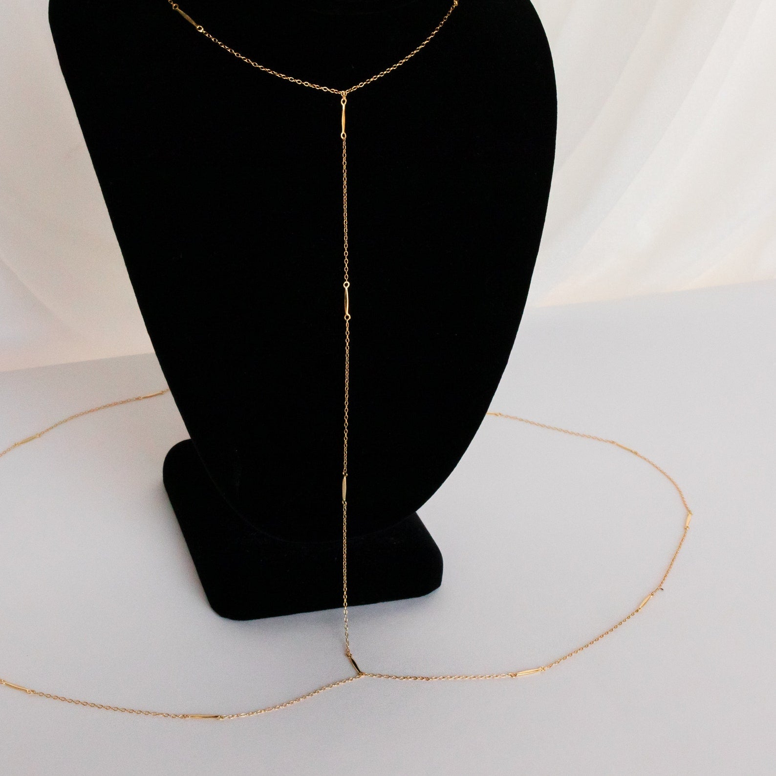 The Kara Body Chain, shown on a black velvet stand with white draped fabric, is the perfect trendy jewelry piece to elevate your summer style.