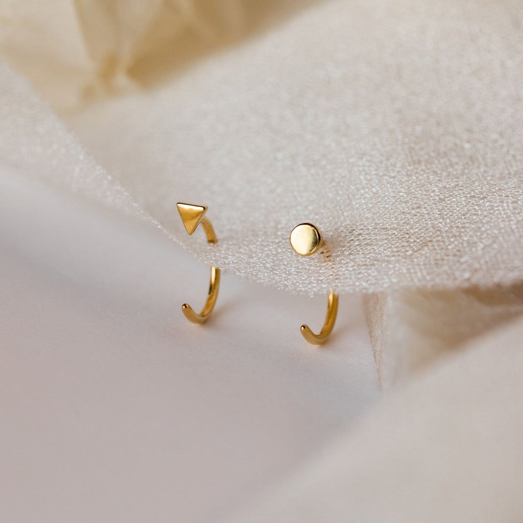 Two gold open huggies—one with a tiny triangle, one with a circle—displayed on beige fabric. The Tiny Circle & Triangle Huggies are a chic pair of handmade, mismatched earrings that add modern flair to any earring stack.