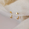 Two gold open huggies—one with a tiny triangle, one with a circle—displayed on beige fabric. The Tiny Circle & Triangle Huggies are a chic pair of handmade, mismatched earrings that add modern flair to any earring stack.