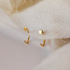 Two gold open huggies—one with a tiny triangle, one with a circle—displayed on beige fabric. The Tiny Circle & Triangle Huggies are a chic pair of handmade, mismatched earrings that add modern flair to any earring stack.