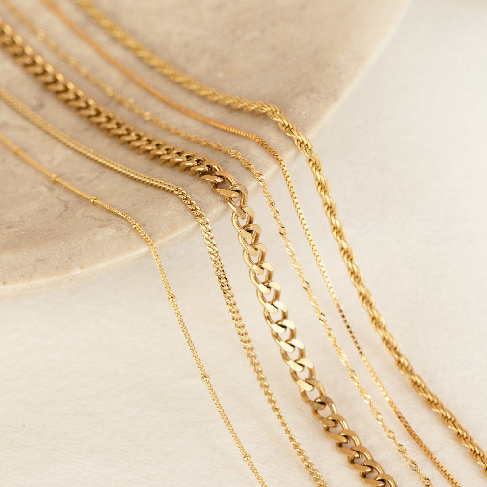 A close-up of various essential gold necklace chains laid out on a neutral-toned stone surface. The image features multiple styles including curb, rope, figaro, and box chains, showcasing their unique textures and link patterns.
