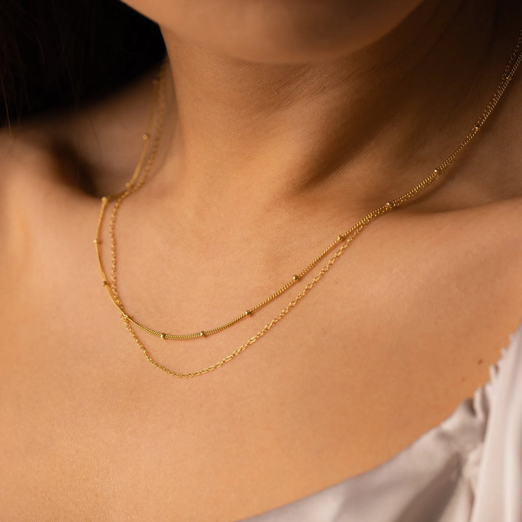 A close-up of a woman in a white satin top wearing two delicate gold layered necklaces, featuring the Beaded Duo Chain Necklace.