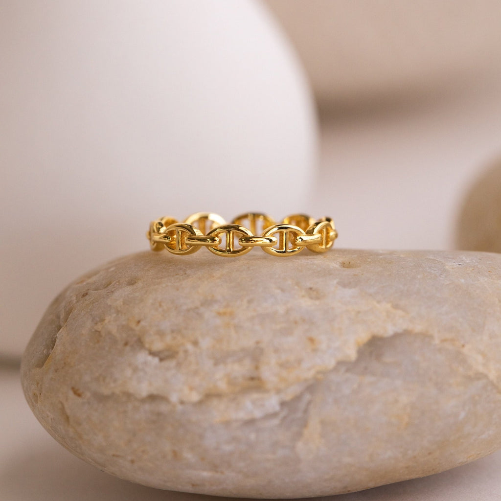 The Reese Link Ring, a minimalist gold chain design, is displayed atop a smooth beige stone against a neutral background.