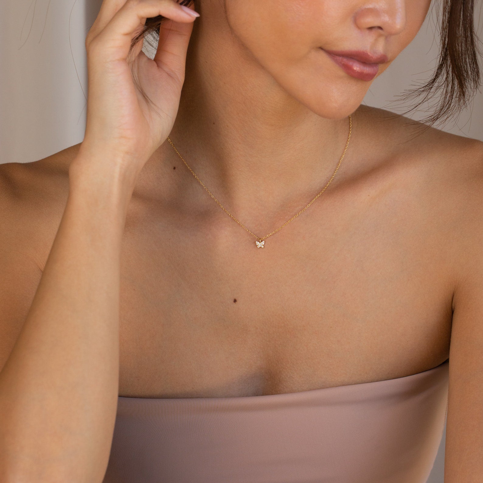 A woman wears the Mandy Pave Butterfly Necklace, featuring gold and diamond details, paired with a strapless beige top as she touches her ear.
