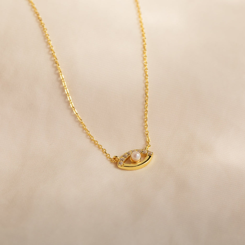 The Amaryllis Pave Evil Eye Necklace in 18K Gold features an eye-shaped pendant with a central pearl and sparkling stones, beautifully displayed on a soft beige background.