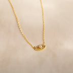 The Amaryllis Pave Evil Eye Necklace in 18K Gold features an eye-shaped pendant with a central pearl and sparkling stones, beautifully displayed on a soft beige background.