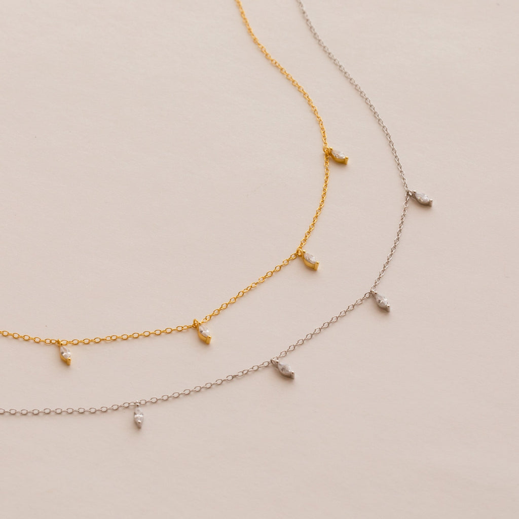 The Marquise Station Necklace, in both gold and silver, features a dainty chain adorned with small marquise-shaped diamond charms and is elegantly displayed on a light background.