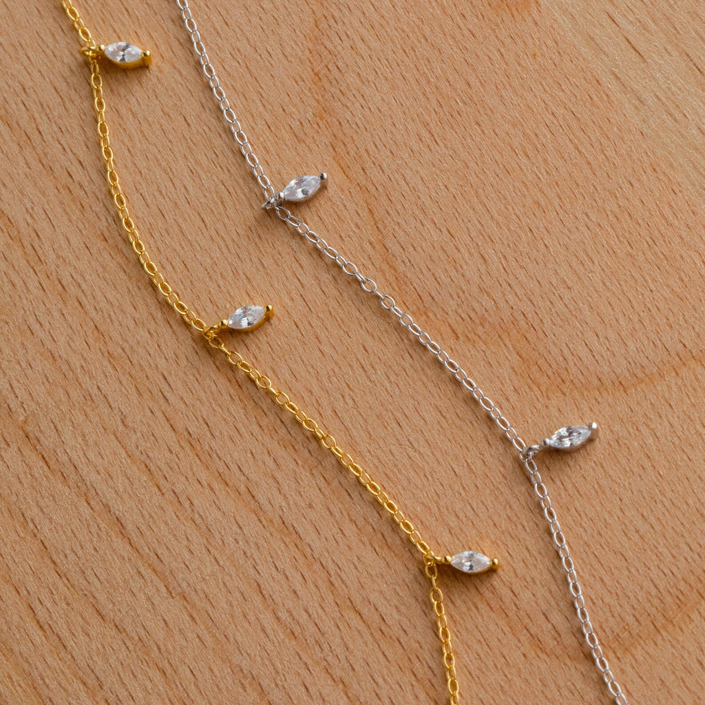 The Marquise Station Necklace features dainty gold and silver chains adorned with small marquise-shaped crystals, displayed on a light wooden surface.