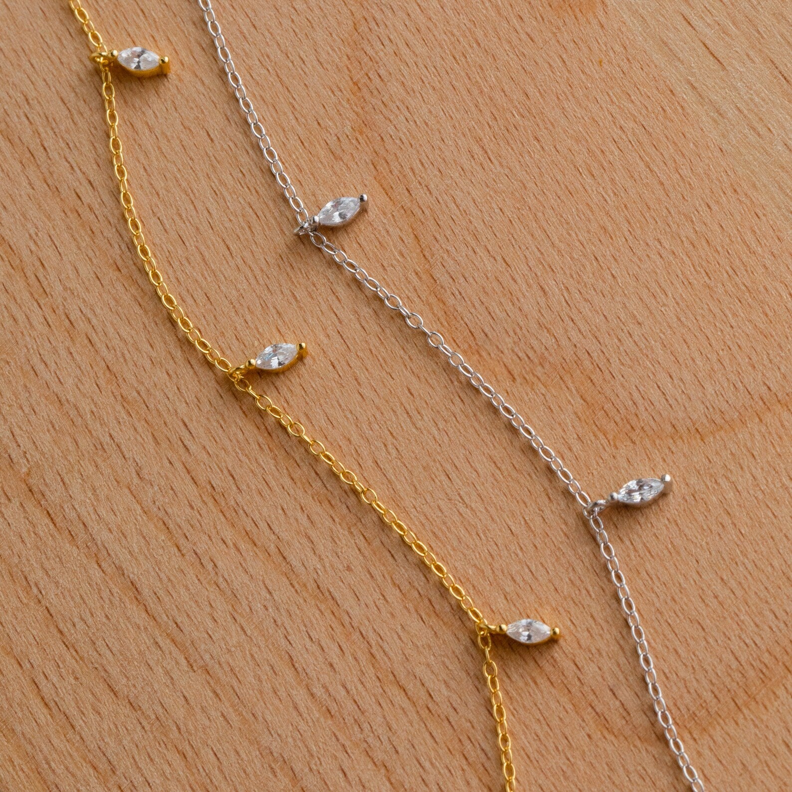 The Marquise Station Necklace features dainty gold and silver chains adorned with small marquise-shaped crystals, displayed on a light wooden surface.