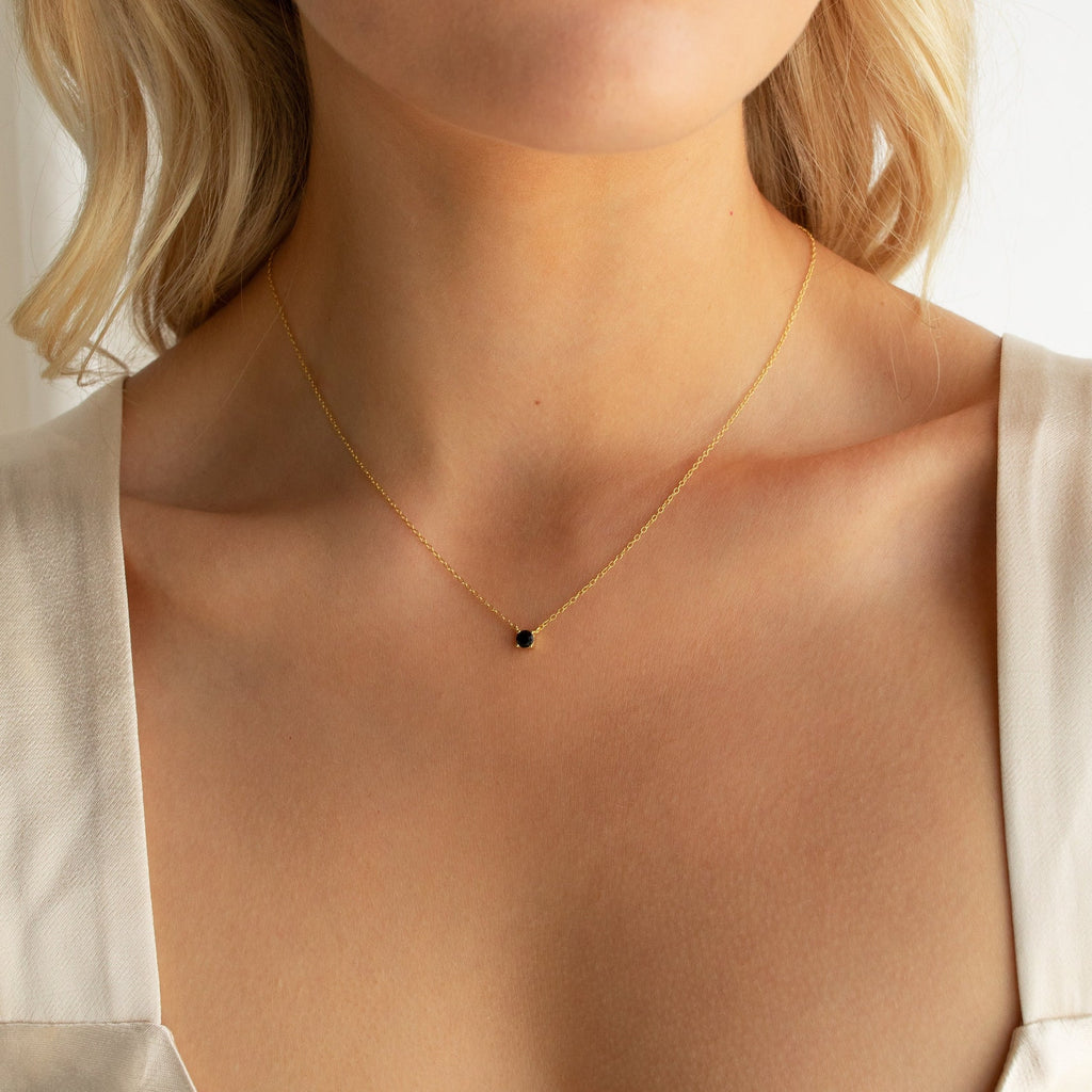 A woman is shown from the shoulders up wearing the Onyx Dainty Diamond Necklace, a vintage-inspired gold chain featuring a small black stone pendant.