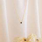 The Onyx Dainty Diamond Necklace, featuring a delicate black stone pendant, is beautifully displayed on soft white fabric with beige flowers—an ideal choice for those wanting a vintage-inspired look.
