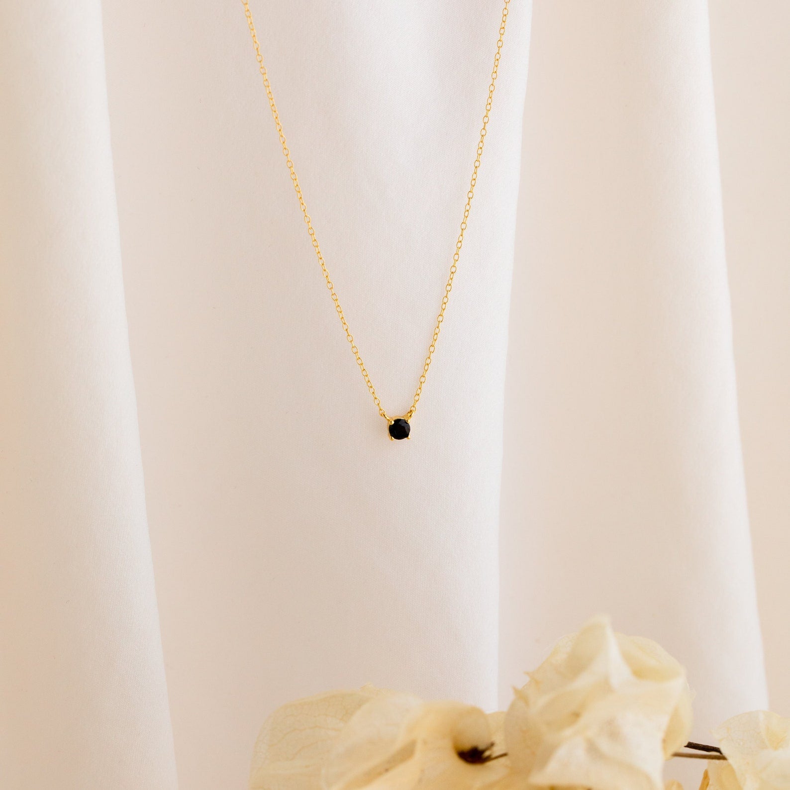 The Onyx Dainty Diamond Necklace, featuring a delicate black stone pendant, is beautifully displayed on soft white fabric with beige flowers—an ideal choice for those wanting a vintage-inspired look.