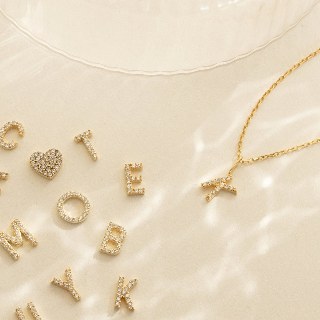 Gold letter and heart charms are scattered on a light surface; to the right lies the Dangling Pave Initial Necklace with a gold K pendant—an elegant gift for women.