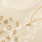Gold letter and heart charms are scattered on a light surface; to the right lies the Dangling Pave Initial Necklace with a gold K pendant—an elegant gift for women.