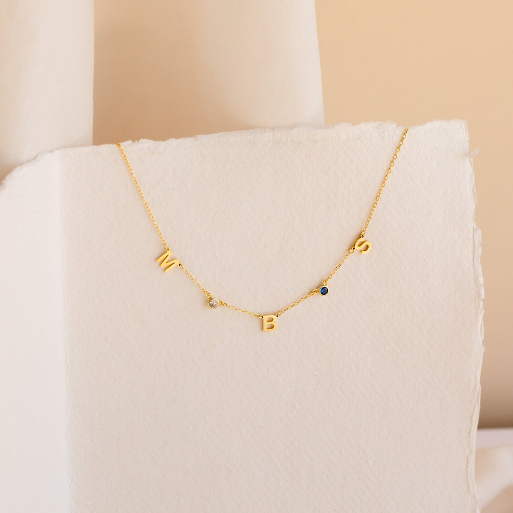 The Alice Letter Birthstone Necklace features gold W, B, S charms and two sparkling gemstones on a white textured background—a unique personalized piece and thoughtful gift for mom.