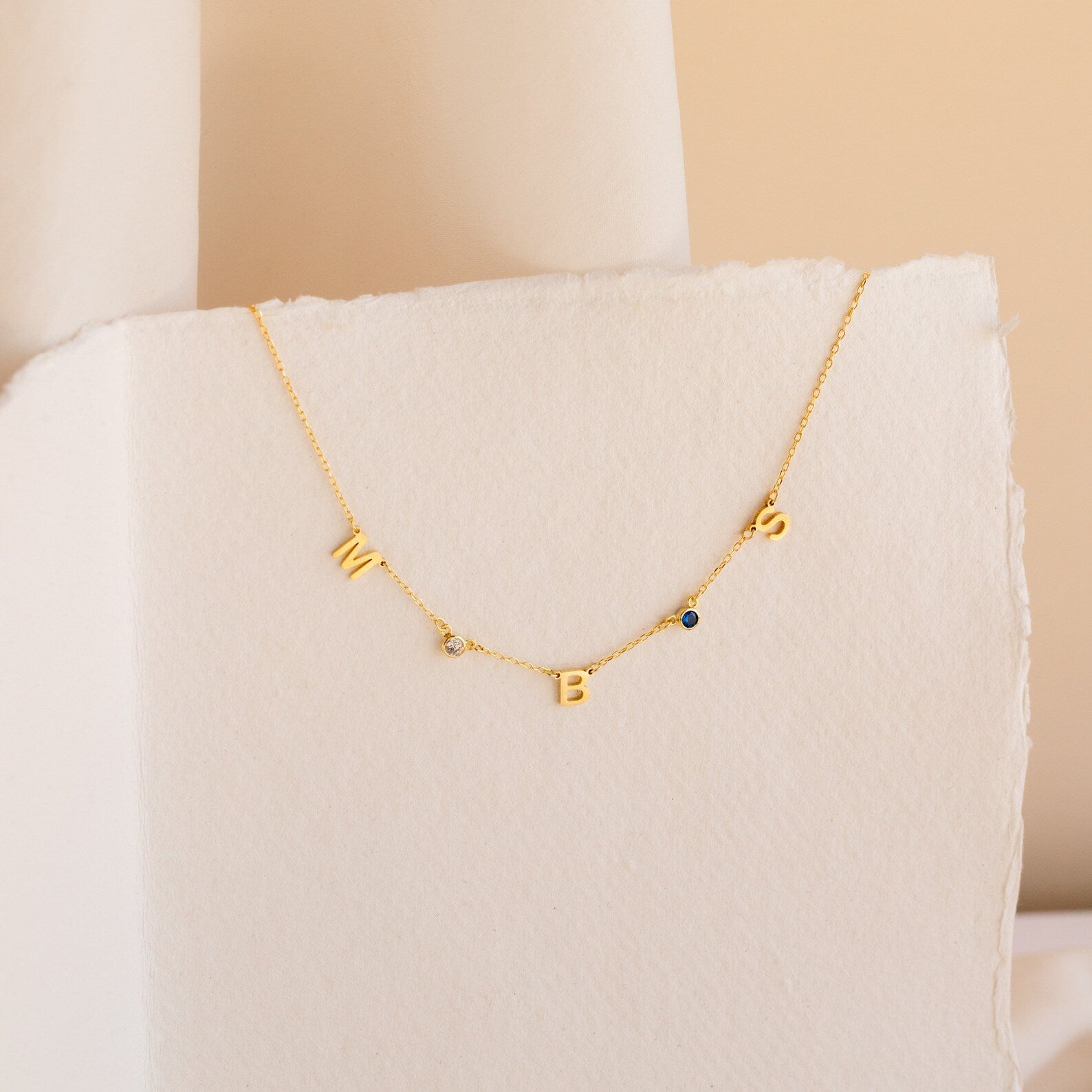 The Alice Letter Birthstone Necklace features gold W, B, S charms and two sparkling gemstones on a white textured background—a unique personalized piece and thoughtful gift for mom.