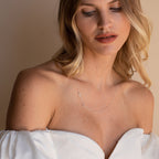 Woman in an off-shoulder white top wears the delicate Sideways Initial Birthstone Necklace, glancing down in soft light—a thoughtful gift for Mom.