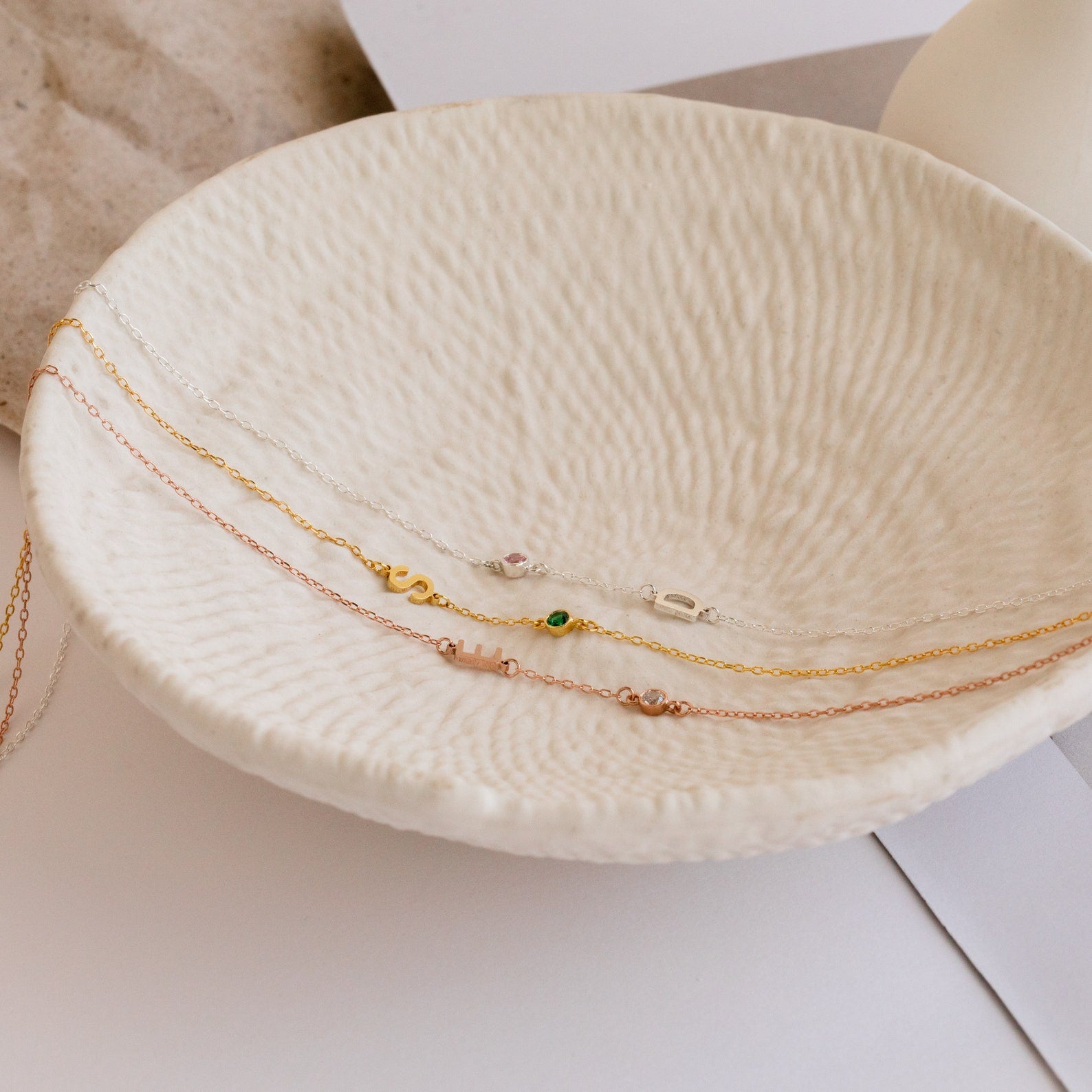 Three delicate gold, silver, and rose gold chain bracelets rest on a cream ceramic dish—an elegant accessory and a thoughtful gift for Mom to pair with her favorite Sideways Initial Birthstone Necklace.