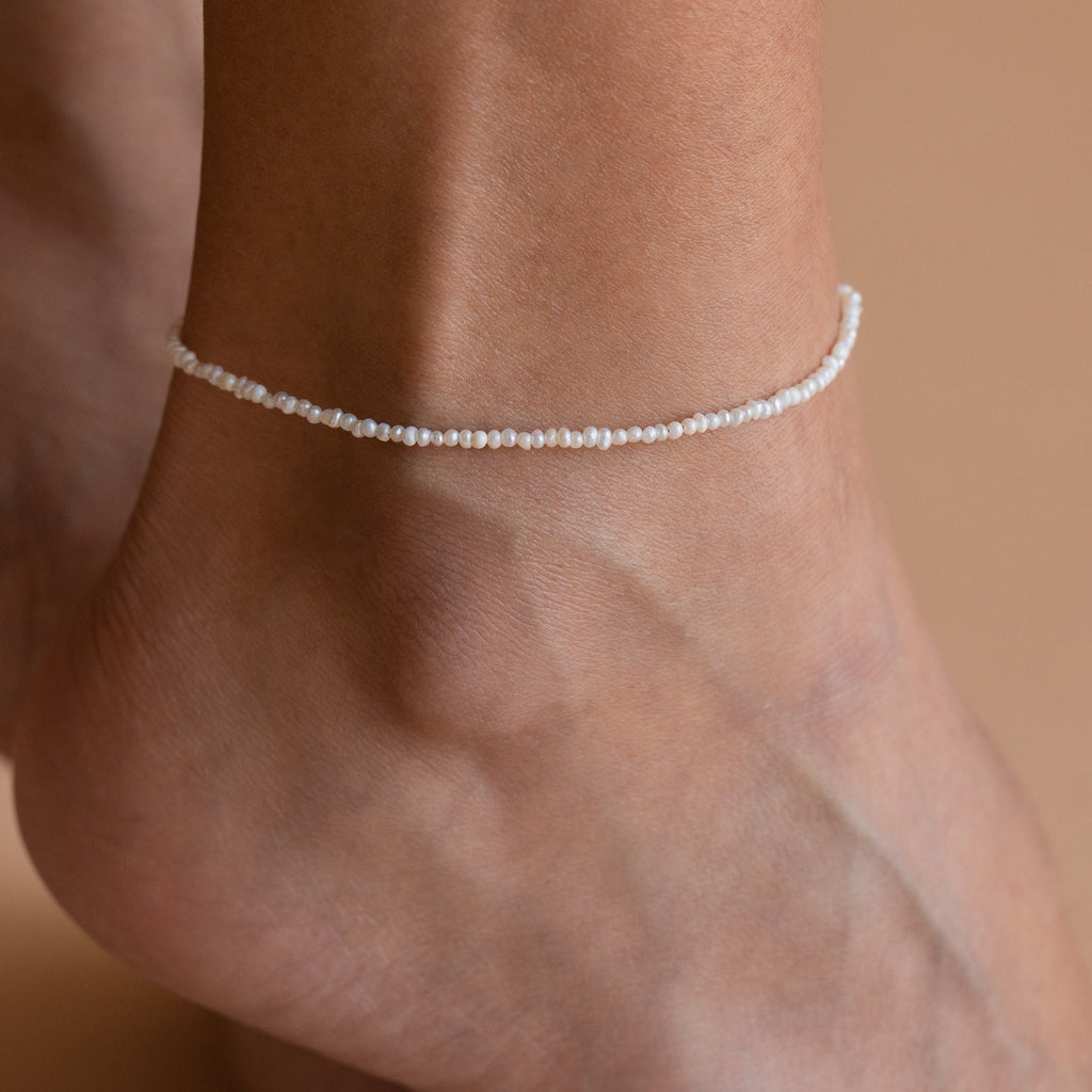 A foot wearing the Brinley Pearl Anklet, which is composed of dainty freshwater pearls strung together, shimmering in the light.