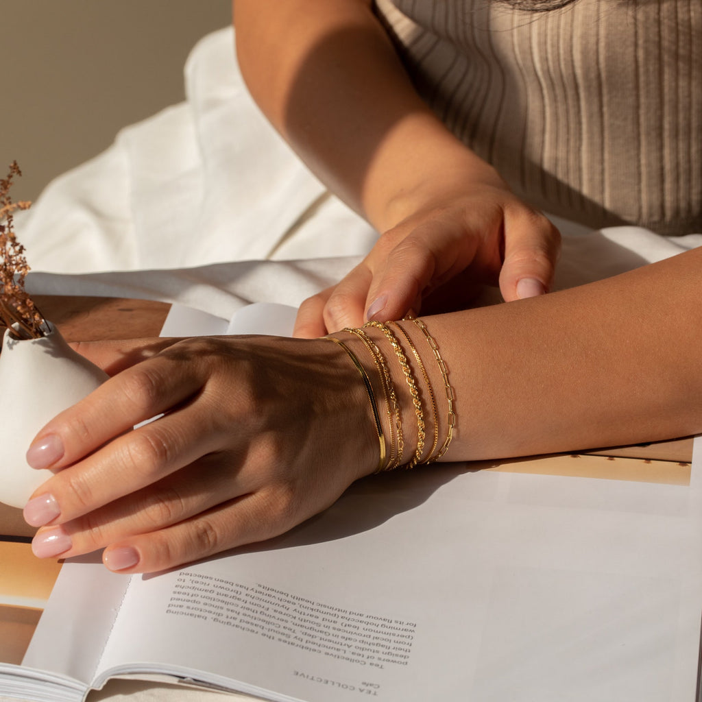 A woman’s hand, adorned with Essential Bracelet Chains, rests on an open magazine in soft natural light.