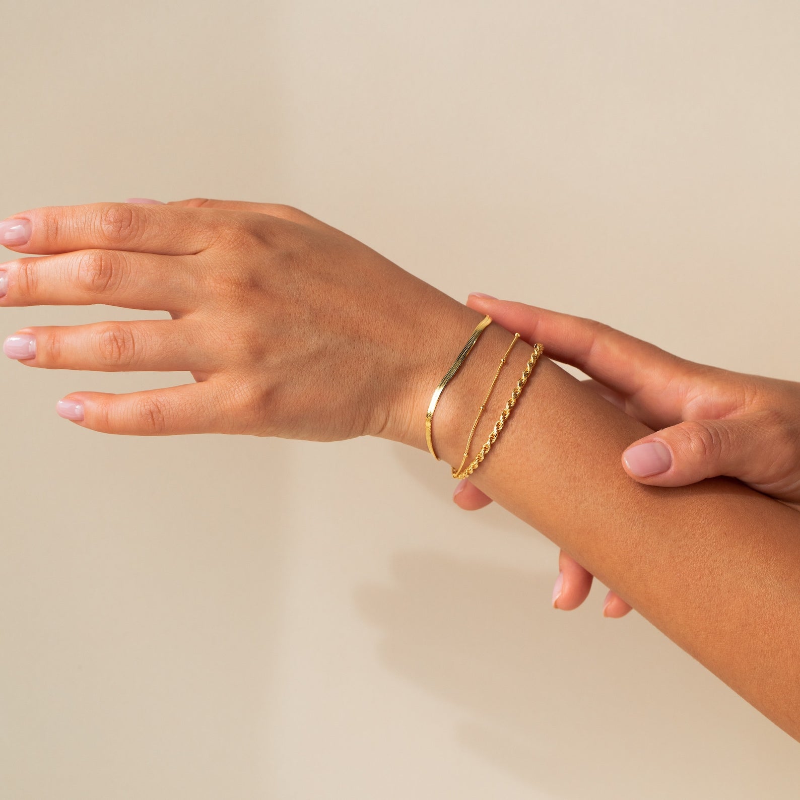 A hand wears two Essential Bracelet Chains in polished gold against a neutral background.
