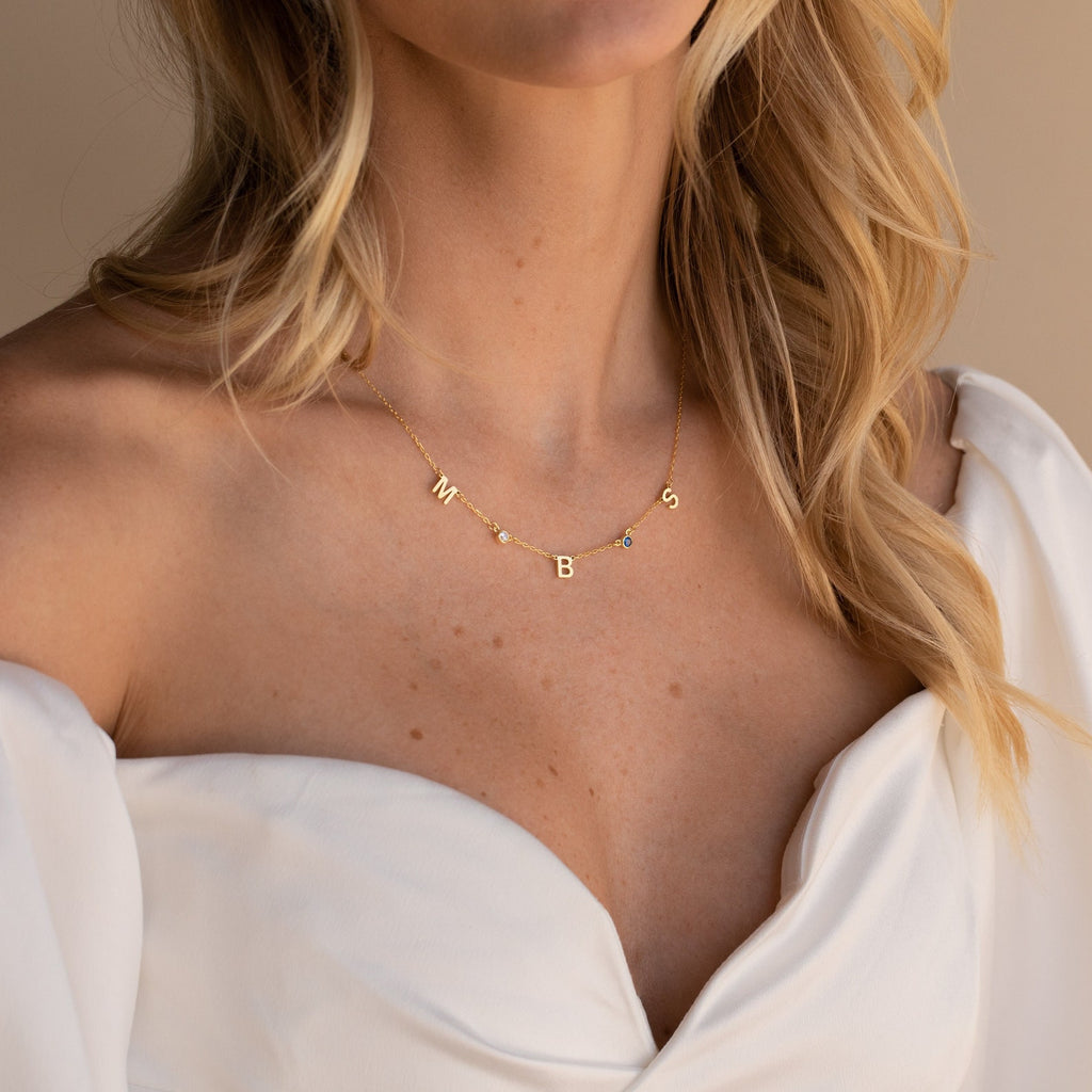 A woman in a white off-shoulder dress wears the Alice Letter Birthstone Necklace with gold M, B, and S charms—a personalized and thoughtful gift for mom.