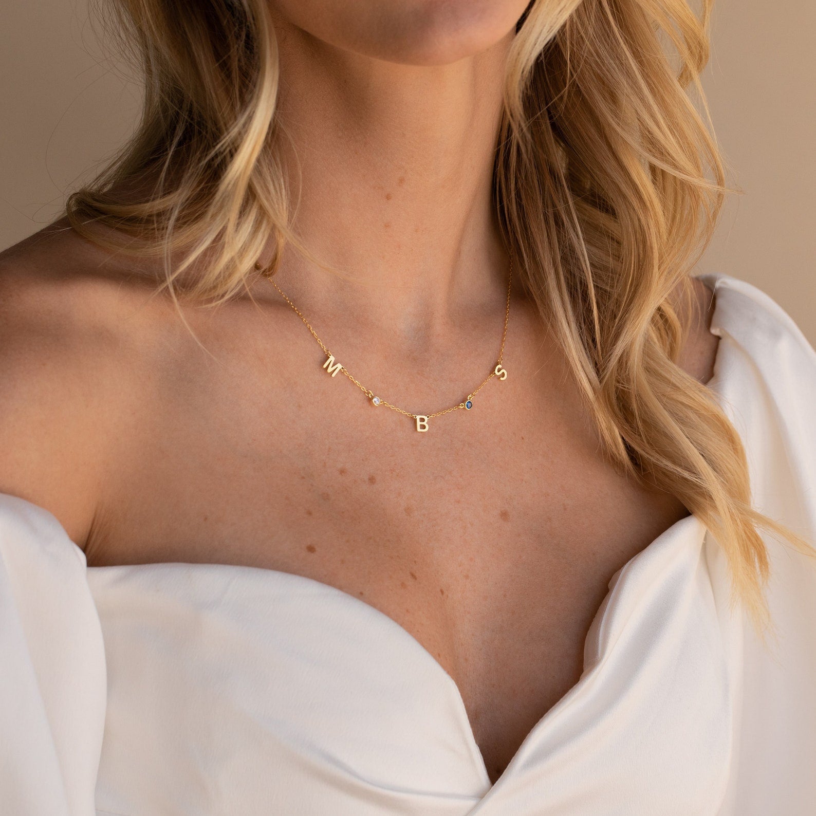 A woman in a white off-shoulder dress wears the Alice Letter Birthstone Necklace with gold M, B, and S charms—a personalized and thoughtful gift for mom.