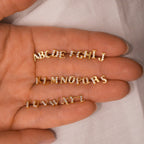A hand displays Pave Initial Studs—gold, diamond-studded alphabet charms in three rows on fabric—ideal for a personalized gift or creating custom initial earrings.