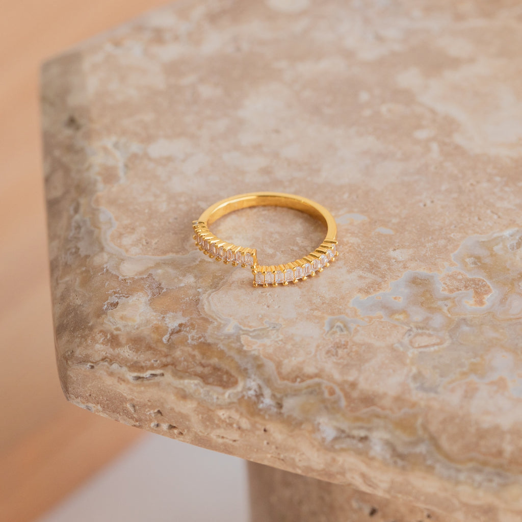 The Elora Baguette Wrap Ring, featuring a V-shaped row of diamond crystal baguettes, rests on a marble surface.