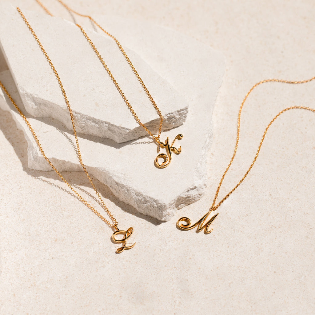Three gold necklaces with cursive letter pendants. The necklaces are on a white surface. The pendants are the letters K, M, and L.