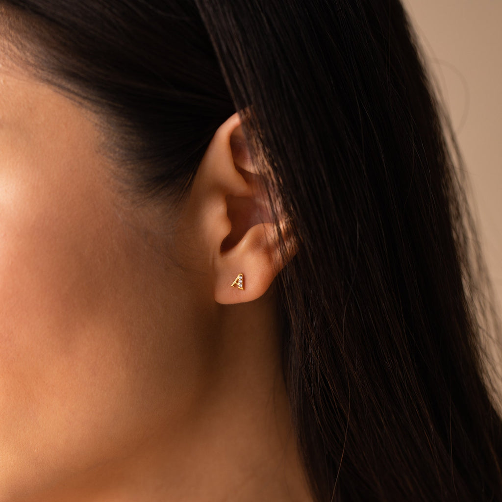 Close-up of a woman's ear with brown hair tucked back, showcasing Pave Initial Studs—small gold triangular earrings that make a perfect personalized gift.