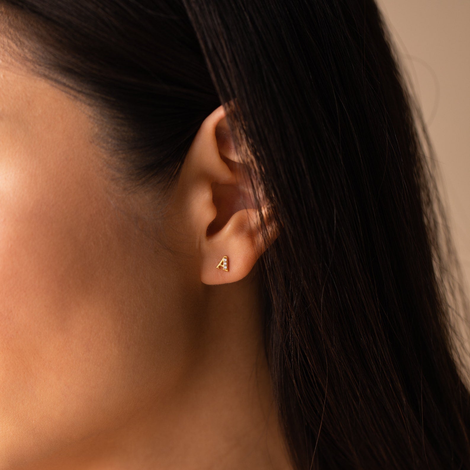 Close-up of a woman's ear with brown hair tucked back, showcasing Pave Initial Studs—small gold triangular earrings that make a perfect personalized gift.