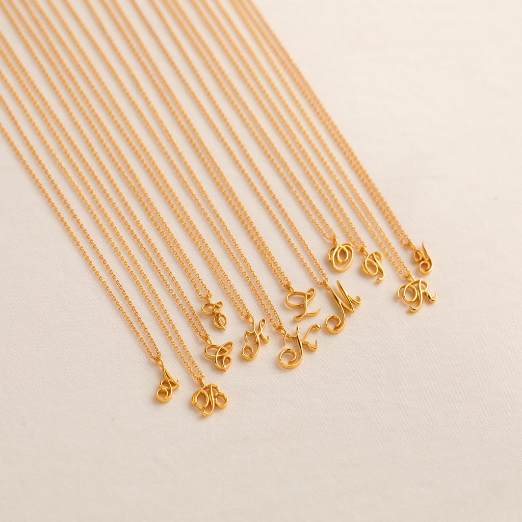 The Davina Letter Necklace features elegant gold cursive initial pendants, beautifully arranged on a light beige background—an ideal romantic gift for someone special.