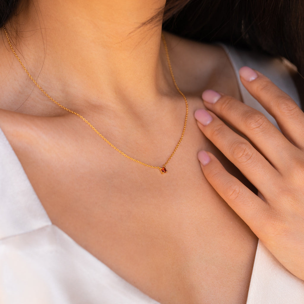 Close-up of a woman wearing the Dainty Birthstone Necklace featuring a small red pendant, touching her collarbone—a perfect choice for lovers of meaningful jewelry.