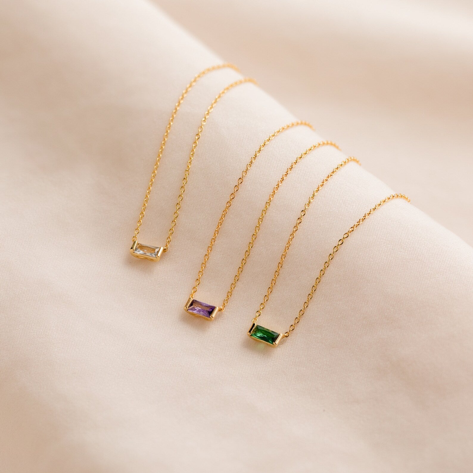 Emery Birthstone Necklaces displayed on a soft fabric background, featuring clear diamond, purple amethyst, and green emerald rectangular stones.
