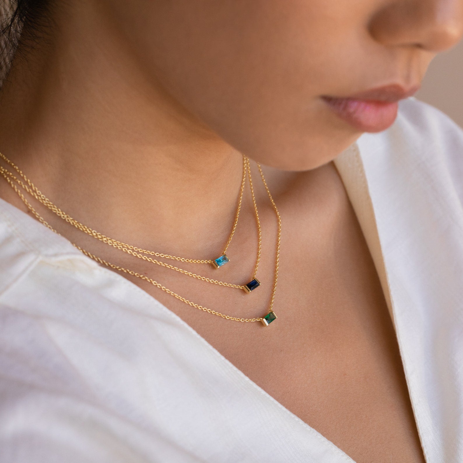A close-up of model wearing three layered Emery Birthstone Necklaces in gold, each with a different rectangular gemstone in blue zircon, sapphire, and emerald