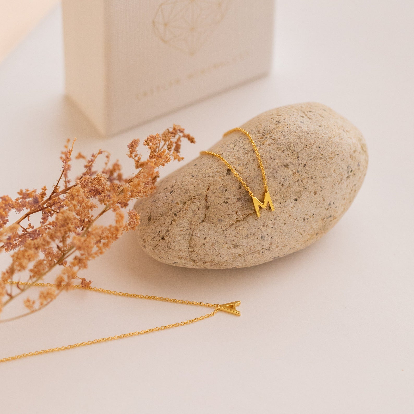 Dainty Initial Necklace: Customizable gold necklaces—choose an "M" on stone or an "A" on cream—styled with dried foliage. A thoughtful Christmas gift.