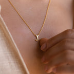A close-up of a woman's chest wearing a delicate gold chain necklace with a polished gold letter 'J' pendant. The pendant rests at the center of her chest.