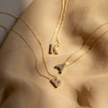 Three Floating Pave Initial Necklaces with diamond-encrusted K, A, and B letter pendants rest on beige fabric—an elegant display of personalized jewelry.