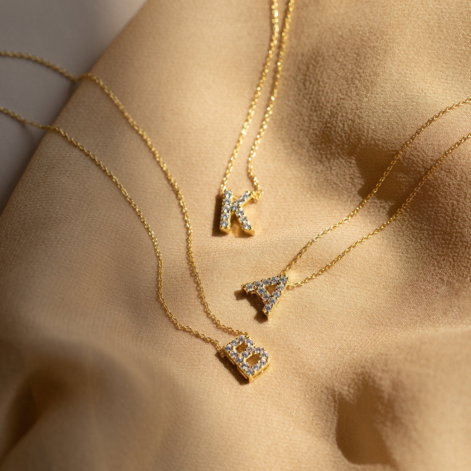 Three Floating Pave Initial Necklaces with diamond-encrusted K, A, and B letter pendants rest on beige fabric—an elegant display of personalized jewelry.