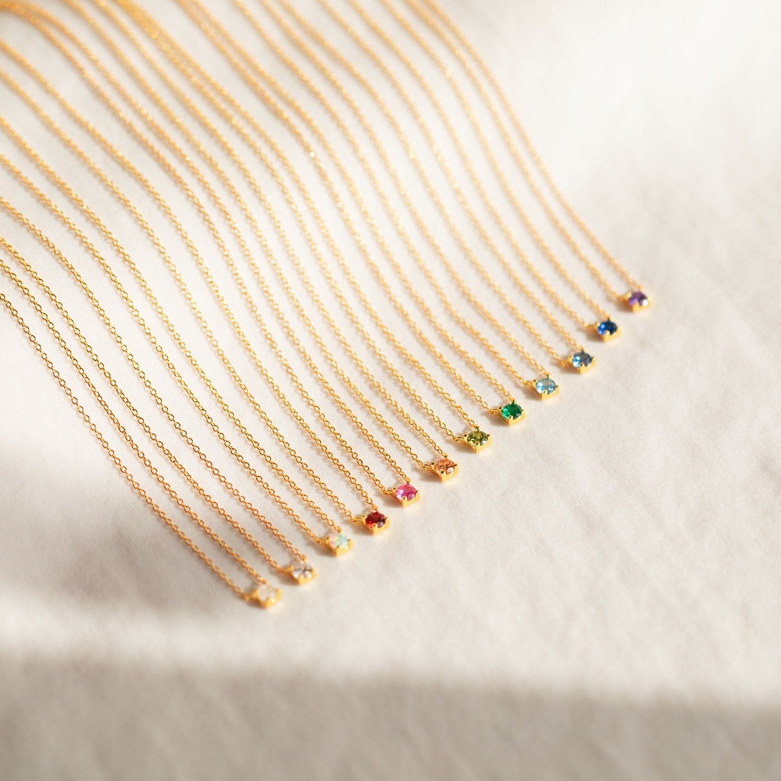 Several Dainty Birthstone Necklaces featuring small, colorful gemstone pendants are arranged in a neat row on a white surface, creating a meaningful jewelry display.